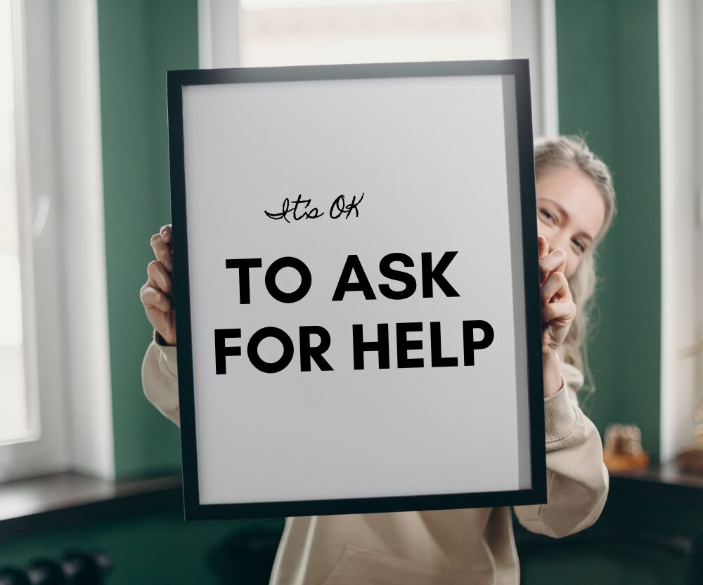 Woman holding a sign stating 'It's Ok to ask for Help', reinforcing the message of our FAQ section.
